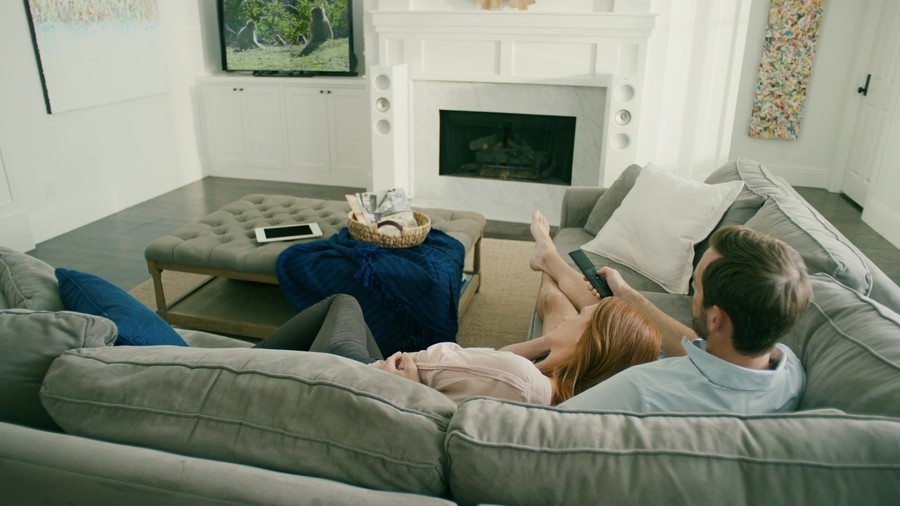 Couple sits on the couch watching TV, changing channels with a Savant remote control